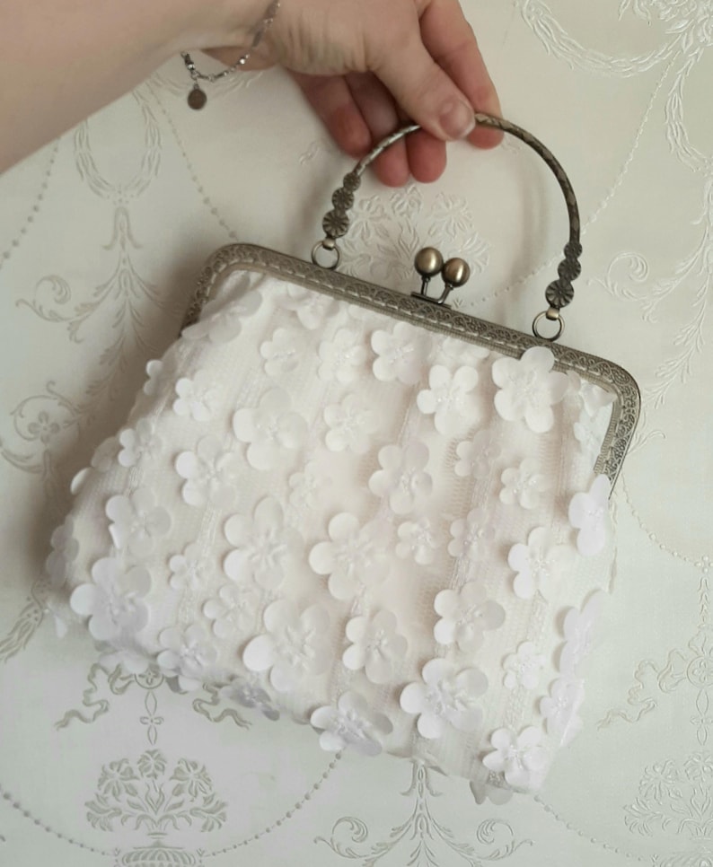 May include: A white clutch purse with a silver frame and a floral design. The purse has a top handle.