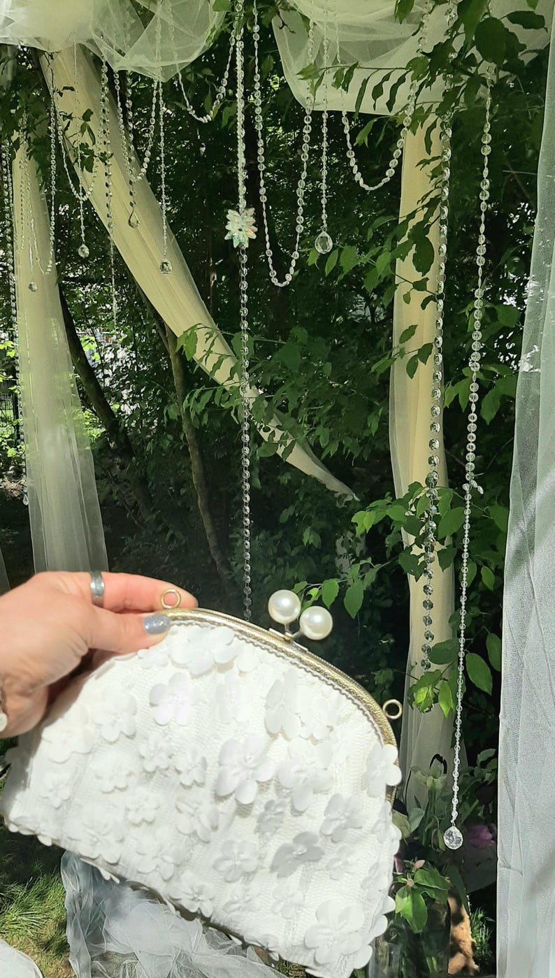May include: A white clutch purse with a floral design and a gold clasp. The purse is decorated with pearl-like embellishments.