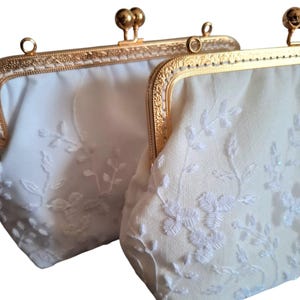May include: Two off-white, vintage-style purses with gold-colored metal frames. The purses feature delicate white floral embroidery and a kiss-lock clasp. The frames have a decorative, ornate design. The purses are likely for special occasions.