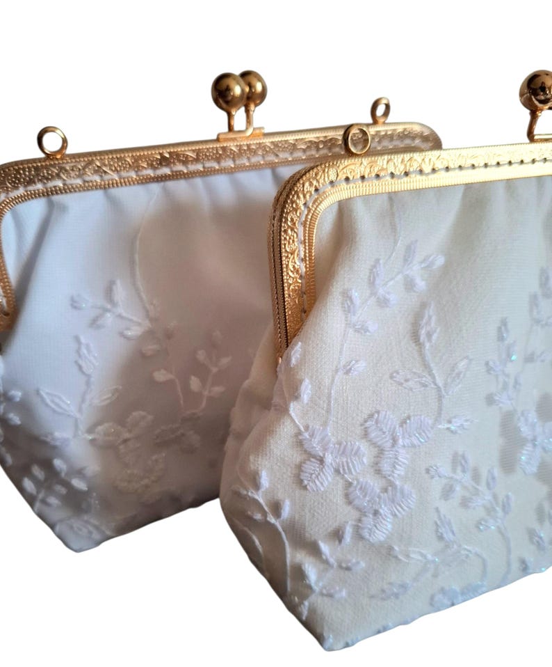 May include: Two off-white, vintage-style purses with gold-colored metal frames. The purses feature delicate white floral embroidery and a kiss-lock clasp. The frames have a decorative, ornate design. The purses are likely for special occasions.