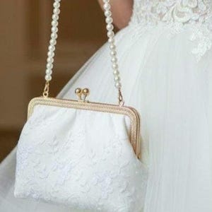 Weiße Brauttasche mit einer perlenbesetzten Kette und einem goldfarbenen Verschluss. Die Tasche hat ein gesticktes Blumenmuster und eine rechteckige Form. Die Tasche wird vor einem weißen Kleid gehalten, was auf eine Hochzeit hindeutet.