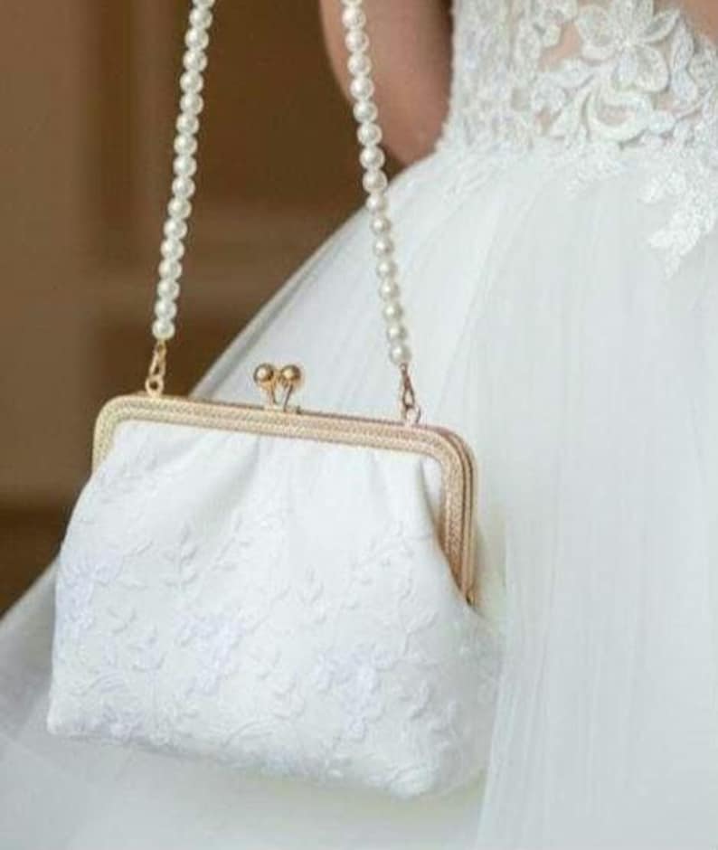 Weiße Brauttasche mit einer perlenbesetzten Kette und einem goldfarbenen Verschluss. Die Tasche hat ein gesticktes Blumenmuster und eine rechteckige Form. Die Tasche wird vor einem weißen Kleid gehalten, was auf eine Hochzeit hindeutet.