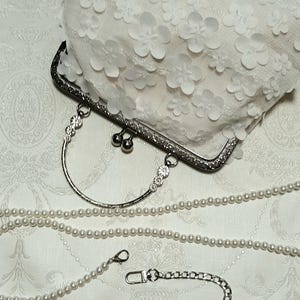 May include: A white clutch purse with a floral design and a silver clasp. The purse comes with a silver chain strap and a pearl chain strap.