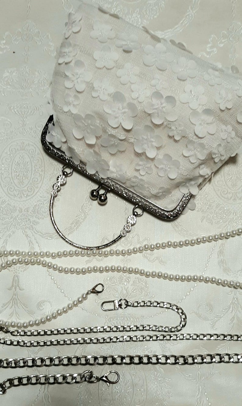 May include: A white clutch purse with a floral design and a silver clasp. The purse comes with a silver chain strap and a pearl chain strap.