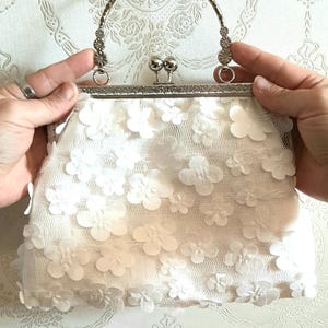 May include: A white clutch purse with a silver clasp and handle. The purse is covered in white fabric flowers.