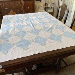 May include: A white tablecloth with a blue and green floral pattern, laid on a dark wood table with chairs.