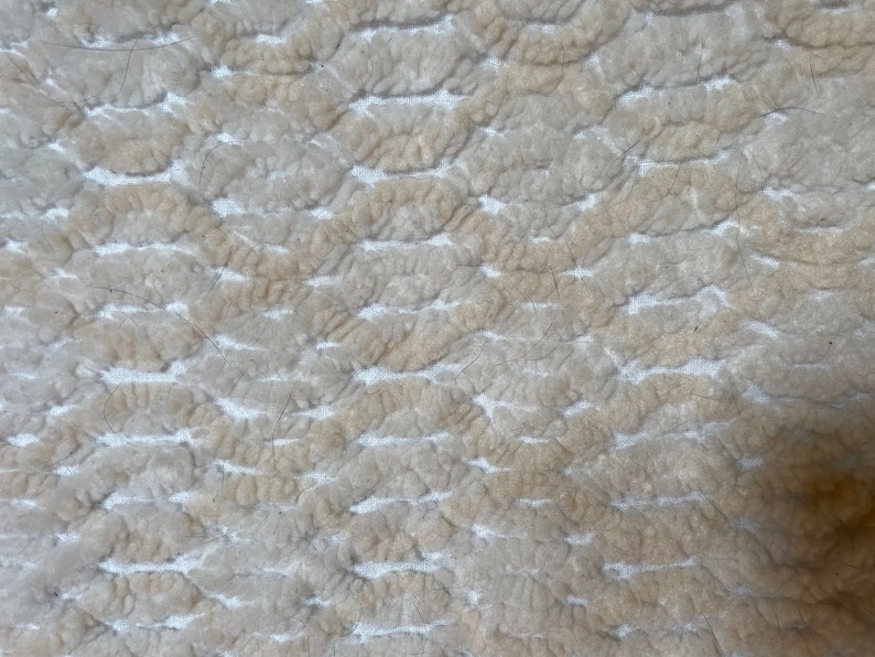 May include: Close-up of a textured, off-white fabric with a pattern of raised, rounded shapes. The fabric has a slightly worn appearance with some loose fibers.