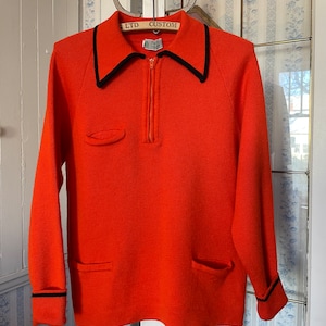 May include: Orange sweater with a black trim around the collar and cuffs. The sweater has a zip up the front and two pockets on the front. The sweater has the text "LTD CUSTOM" on the collar.