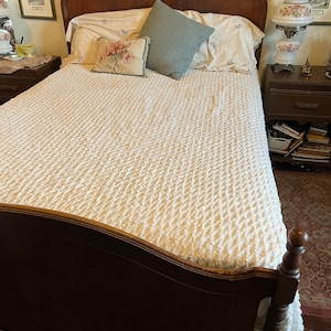 May include: A wooden bed frame with a cream-coloured, textured bedspread. The bed is made with floral patterned pillows and a light blue pillow. A bedside table with a lamp and framed artwork is on the right.