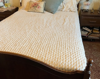 Puede incluir: Una cama de madera con una colcha texturizada de color crema. La cama está hecha con almohadas con estampado floral y una almohada azul claro. Una mesita de noche con una lámpara y obras de arte enmarcadas está a la derecha.