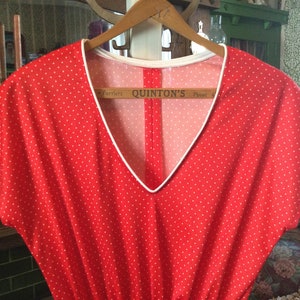 May include: Red sleeveless dress with white polka dots and a white trim around the neckline. The dress is hanging on a wooden hanger. The tag on the hanger reads "Quinton's Phone".