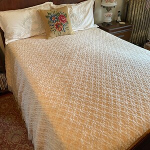 May include: A white bedspread with a textured pattern, featuring a floral embroidered pillow on top of a white pillow. The bedspread has a fringe border.