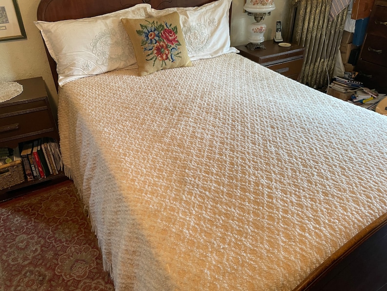 May include: A white bedspread with a textured pattern, featuring a floral embroidered pillow on top of a white pillow. The bedspread has a fringe border.