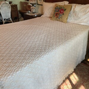 May include: A white bedspread with a textured pattern and fringe trim. The bedspread is on a bed with white pillows and a floral pillow.