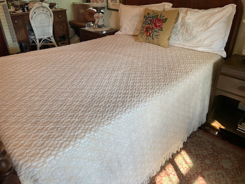 May include: A white bedspread with a textured pattern and fringe trim. The bedspread is on a bed with white pillows and a floral pillow.