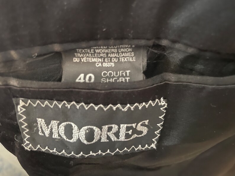 Puede incluir: Una etiqueta negra con texto blanco que dice "MIER CUIMING TEXTILE WORKERS UNION TRAVAILLEURS AMALGAMES DU V&Ecirc;TEMENT ET DU TEXTILE CA 05375 COURT 40 SHORT" y una etiqueta negra con costuras blancas que dice "MOORES".