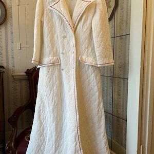 May include: A white quilted dressing gown with red piping. The gown has a long, flowing silhouette and a double-breasted fastening.