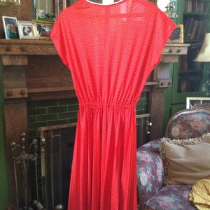 May include: A red dress with white polka dots. The dress has a gathered waist and a slightly flared skirt. It is hanging on a hanger in front of a fireplace.