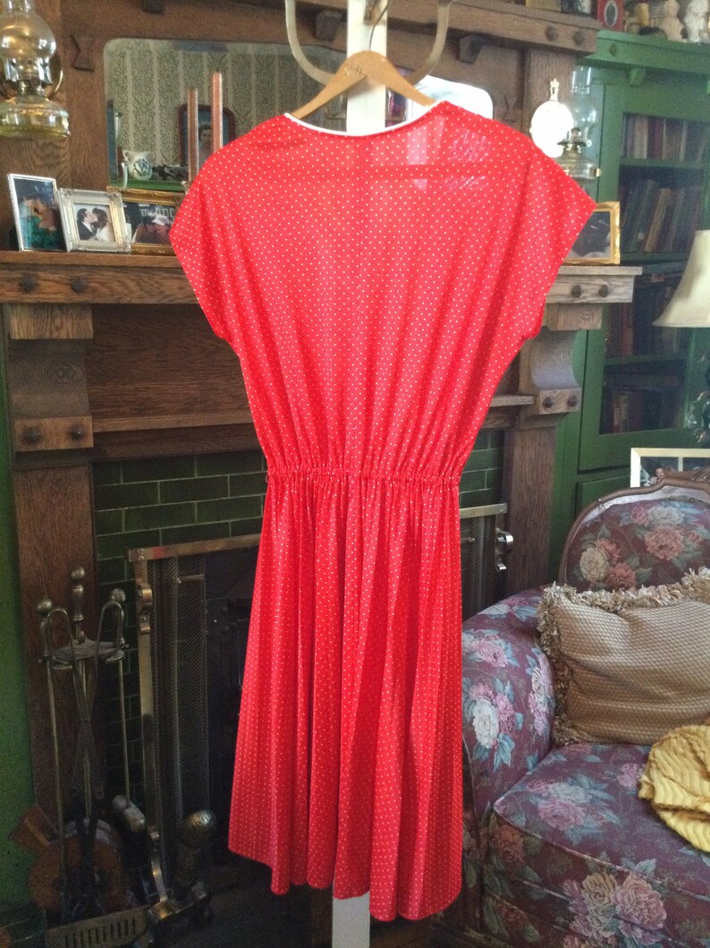 May include: A red dress with white polka dots. The dress has a gathered waist and a slightly flared skirt. It is hanging on a hanger in front of a fireplace.