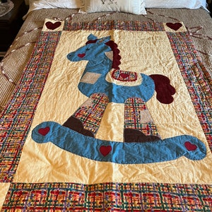 May include: A cream-coloured quilt with a blue rocking horse appliqué. The horse has a brown mane and tail, and red hearts are sewn around the horse and on the quilt's border. The border is a plaid fabric with red, green, yellow, and blue colours.