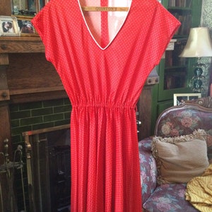 May include: A red dress with white polka dots and a white v-neck collar. The dress has a gathered waist and a flowing skirt. The dress is hanging on a wooden hanger.
