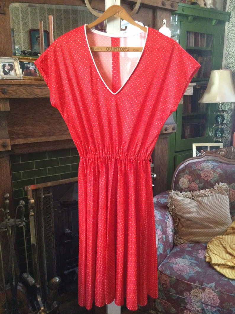 May include: A red dress with white polka dots and a white v-neck collar. The dress has a gathered waist and a flowing skirt. The dress is hanging on a wooden hanger.