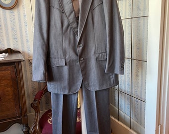 Puede incluir: Una chaqueta de traje gris de rayas finas y pantalones a juego colgados de un soporte blanco. La chaqueta tiene un cierre de un solo botón y un cuello puntiagudo.