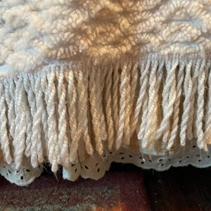 May include: Close-up of a white woven rug with a fringe border. The fringe is made of twisted yarn.