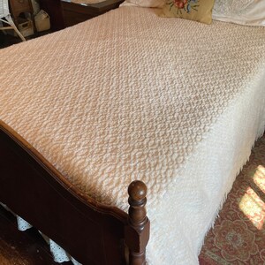 May include: A white bedspread with a textured pattern and fringe. The bedspread is on a wooden bed frame with a dark brown finish.