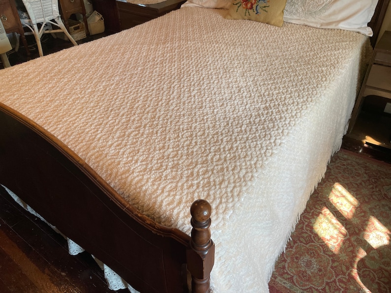 May include: A white bedspread with a textured pattern and fringe. The bedspread is on a wooden bed frame with a dark brown finish.