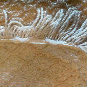 May include: Close-up of a cream-colored rug with a white fringe. The rug has a woven pattern of small, repeating shapes.