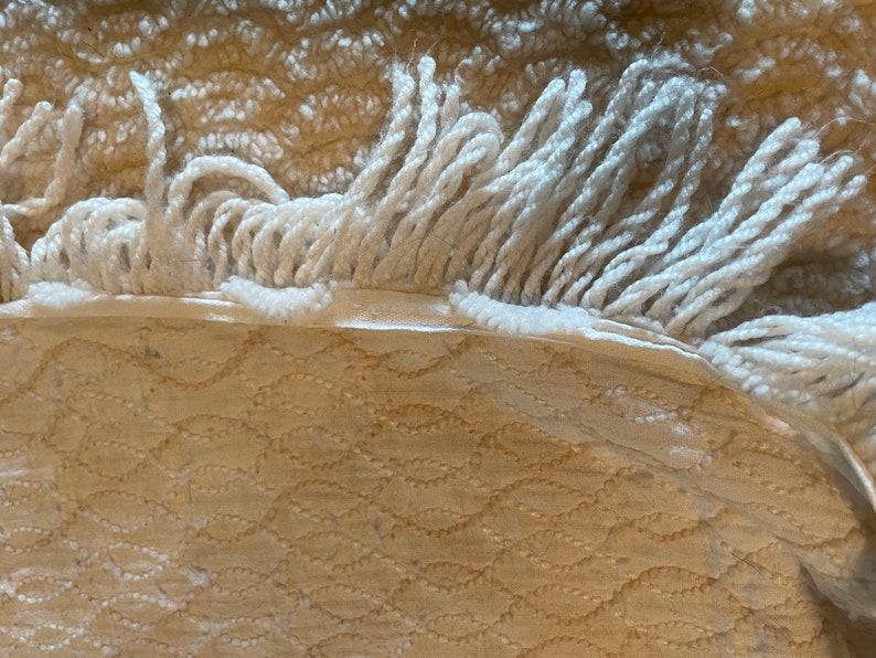 May include: Close-up of a cream-colored rug with a white fringe. The rug has a woven pattern of small, repeating shapes.