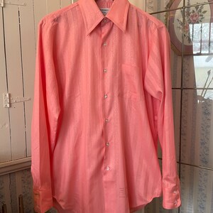 May include: A long-sleeved, pink button-down shirt with a subtle striped pattern. The shirt has a single pocket on the left chest and a brand label on the collar.