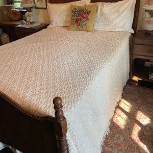 May include: A white bedspread with a textured pattern and fringe trim, on a wooden bed frame with a carved headboard. The bed is made with white pillows and a floral embroidered pillow.