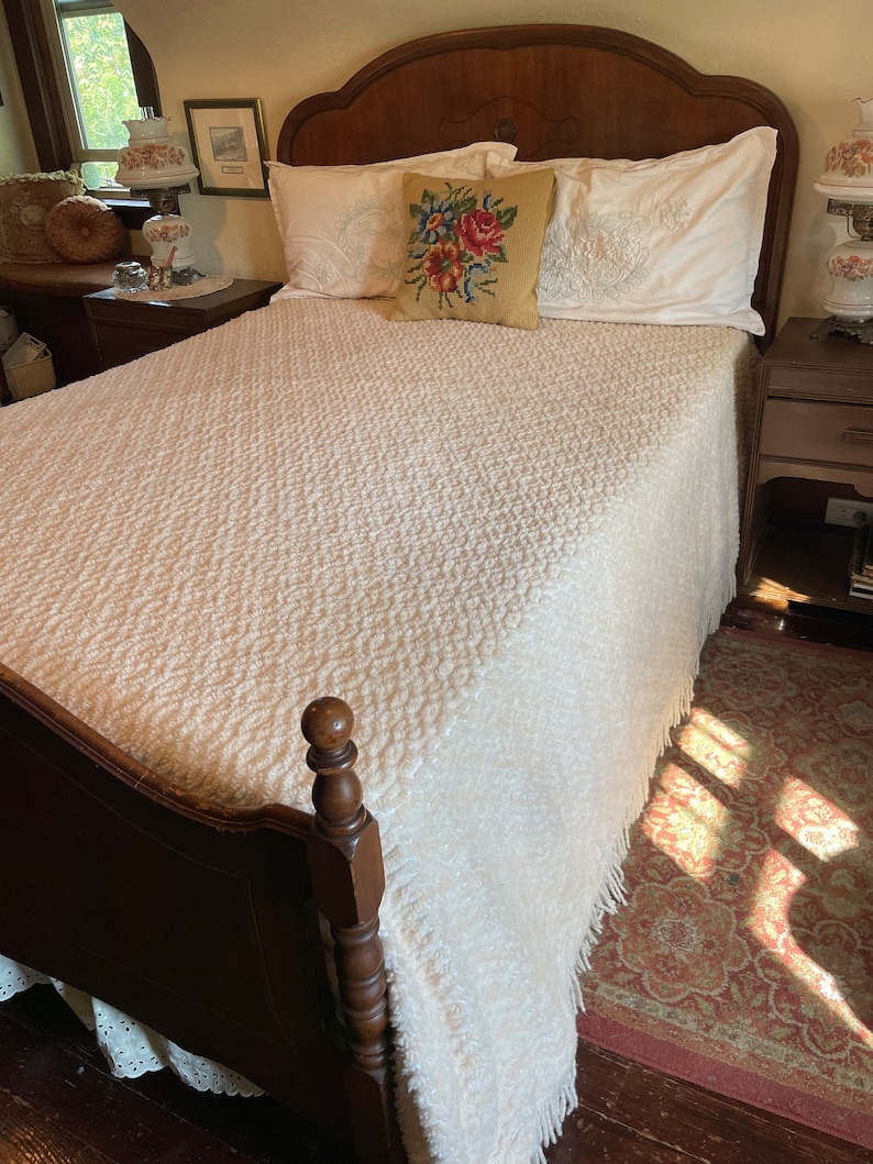 May include: A white bedspread with a textured pattern and fringe trim, on a wooden bed frame with a carved headboard. The bed is made with white pillows and a floral embroidered pillow.
