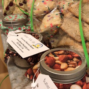 May include: Three glass jars with lids, each containing a different type of gourmet flavored butter. The jars are labeled "Butter Me Up" and feature a cartoon bee logo. The jars are sitting on a countertop with a bag of cookies in the background.