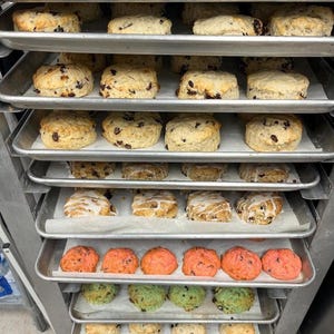 May include: Multiple trays of baked goods, including scones, biscuits, and cookies, are stacked on top of each other. The trays are lined with parchment paper and the baked goods are arranged in rows. The baked goods are various colors, including white, brown, and pink.