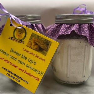May include: Two glass jars with purple fabric lids and a yellow label that reads "Butter Me Up's Lavender Scones Make your own scone(s)! Just add butter and buttermilk www.ButterMeUpNJ.com 732-930-1116".