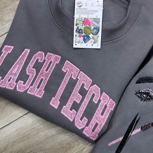 Peut inclure: Sweat-shirt gris avec inscription "LASH TECH" en paillettes roses. Le sweat-shirt a un œil noir et blanc et des outils de cils sur la manche.
