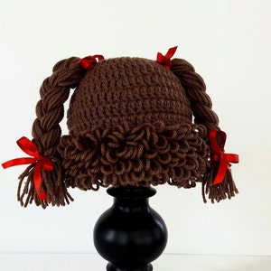 May include: Brown crocheted hat with two braided pigtails and red bows. The hat is displayed on a black pedestal.