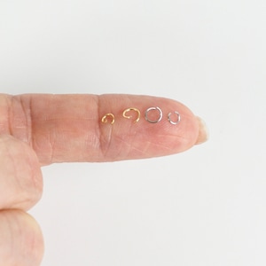 May include: Five jump rings in different sizes, four are gold and one is silver. The rings are displayed on a finger.