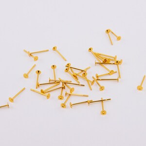 May include: A collection of gold-colored earring posts scattered on a white surface. Each post has a flat, circular disc at one end and a small, straight post extending from the center of the disc. The posts are of varying lengths.