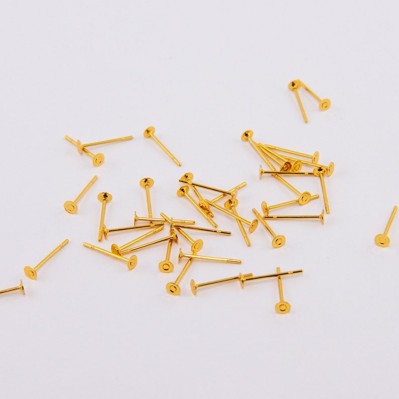May include: A collection of gold-colored earring posts scattered on a white surface. Each post has a flat, circular disc at one end and a small, straight post extending from the center of the disc. The posts are of varying lengths.