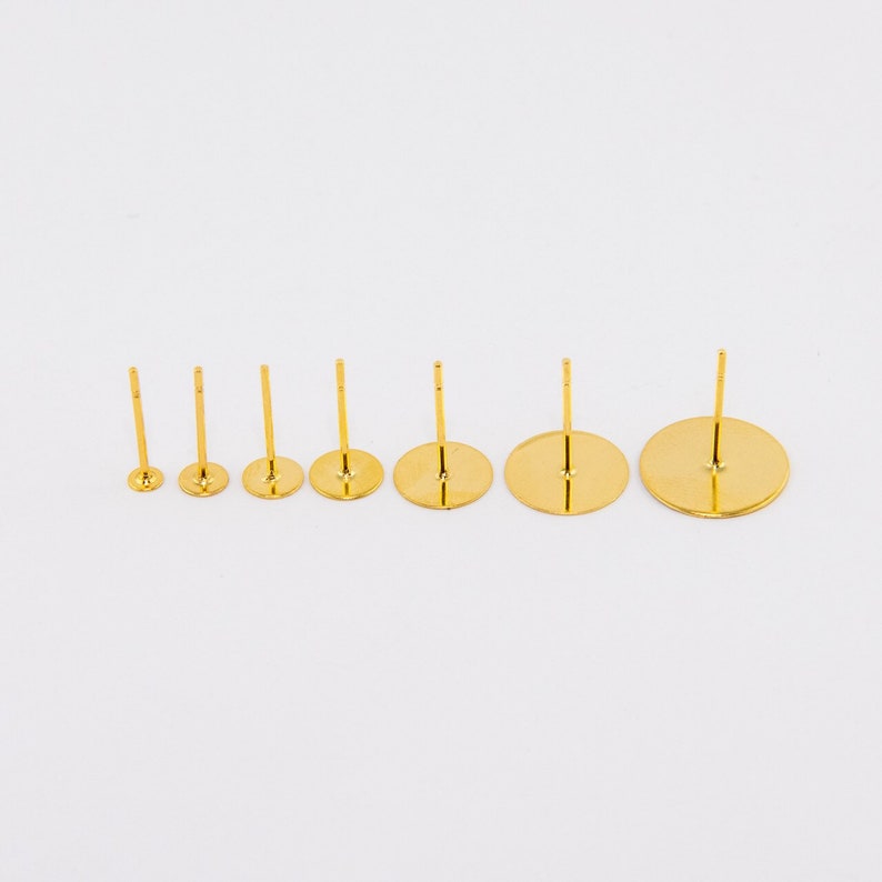 May include: A collection of six gold-colored earring posts with varying disc sizes. Each post has a flat, round base and a vertical post for attaching earrings. The posts are arranged in a row against a white background.