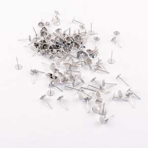 May include: A collection of silver-colored earring posts with flat, round pads and thin, straight posts. The earring findings are scattered on a white surface, with some posts standing upright and others lying flat.