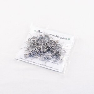 May include: A clear plastic bag filled with silver-colored push pins. The push pins have a flat, circular head and a sharp pin. The bag has text that reads "MF Creative Supplies" and "Plastic packaging is NatureFlex, a 100% compostable plastic."