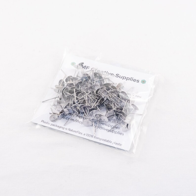 May include: A clear plastic bag filled with silver-colored push pins. The push pins have a flat, circular head and a sharp pin. The bag has text that reads "MF Creative Supplies" and "Plastic packaging is NatureFlex, a 100% compostable plastic."