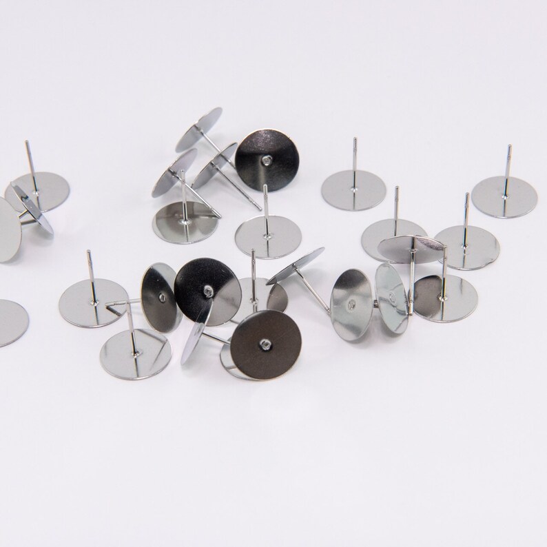May include: A collection of silver-colored earring posts with flat, circular pads and thin, straight posts. The posts are scattered on a white surface, with some slightly angled. The earring findings are for jewelry making.