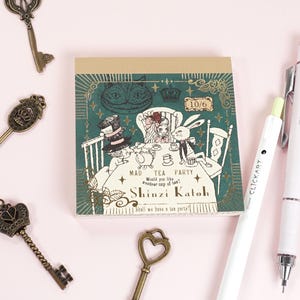 May include: A notepad featuring an Alice in Wonderland Mad Tea Party illustration, with the Cheshire Cat, Mad Hatter, and White Rabbit. The notepad has the text "Shinzi Katoh" and "Mad Tea Party". Decorative keys and pens surround the notepad.