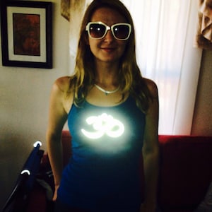 Op de afbeelding: Een vrouw draagt een witte zonnebril en een blauw tanktop met een reflecterend wit Om-symbool op de voorkant.